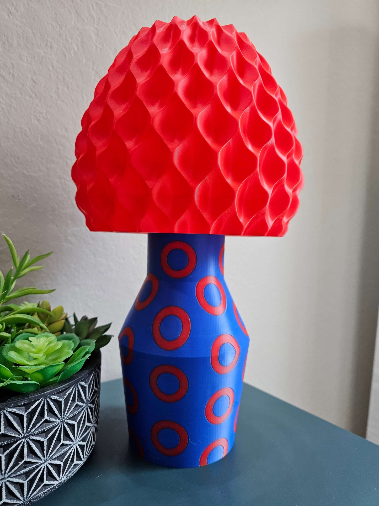 Phishy Donut Lamp with red textured shade and blue base featuring red circles, next to a potted plant on a table.